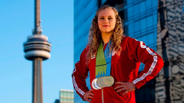 A woman in a red tracksuit poses outdoors with her hands on her hips, with five medals hung around her neck.