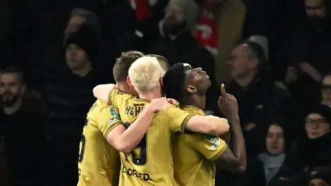 Reuters Crystal Palace's Marc Guehi is looking up to the sky and pointing. Several of his team mates are hugging him from behind. They are all wearing gold jerseys. 