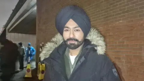 BBC/Naj Modak Tarsem Singh Jassar wearing a blue turban and blue coat. He has a black beard. He is wearing a white t-shirt, grey shirt and grey top. There is a brick wall underneath a bridge behind him. There are two yellow traffic cones and members of the cast and crew stand in the background.