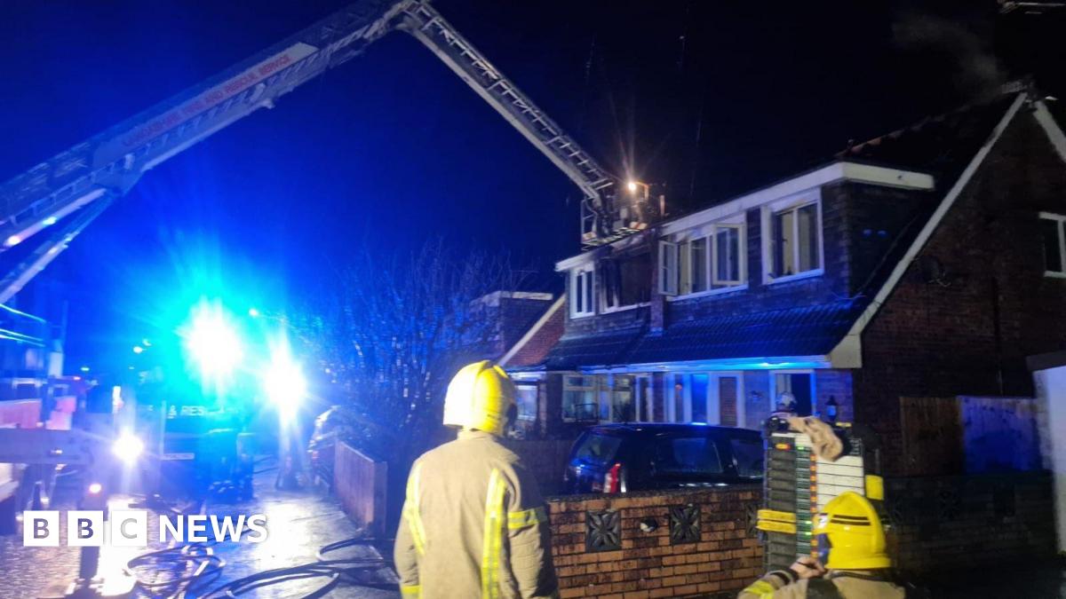 Two firemen in the foreground looking towards a house at night time. There is a crane with platform outside a burnt out window.