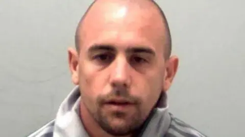 Essex Police Custody picture of Ben Ransome. He has a shaved head and stubble. He looks resigned and is wearing a grey Adidas jumper.