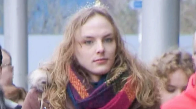 a transgender woman with long brown hair is wearing a brown winter jacket and colourful scart