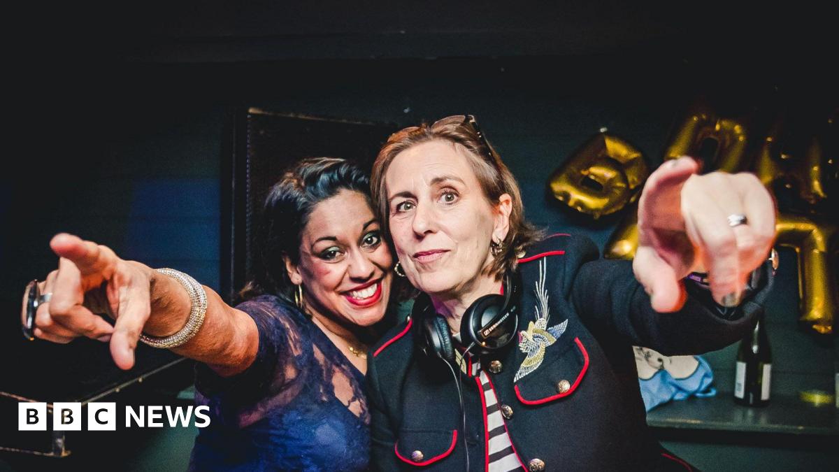 Two women - Aarti Joshi and Kirsty Wark - standing behind the decks in a club. Both are pointing towards the camera, with headphones round the neck of Kirsty Wark.