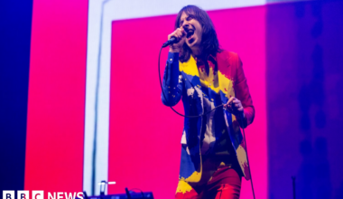 A man wearing a multi coloured jacket, shirt and trousers, sings into a microphone while performing on stage.