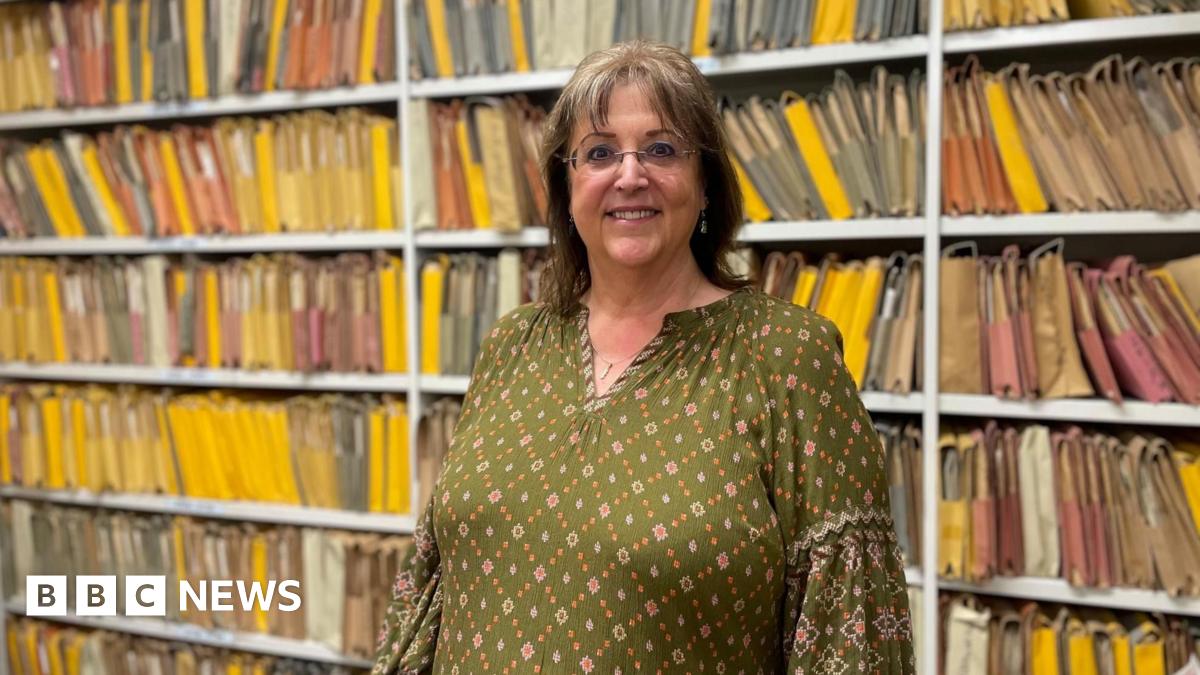 A woman with dark shoulder length hair and glasses smiles for the camera. She is wearing a dark green flowered top in front of a white shelving unit with colourful paper files
