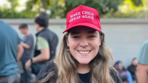 Chilean voter Augustina Trancoso smiles toward the camera. She is wearing a red "Make Chile Great Again" cap. 