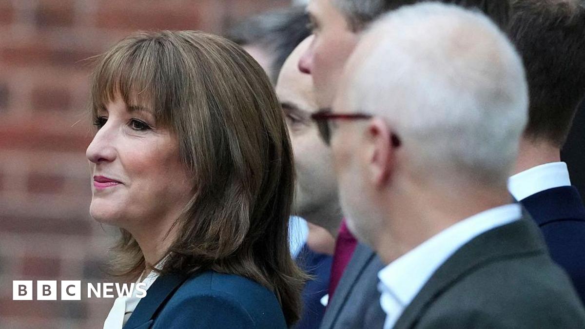 A profile close-up photo of Rachel Reeves, dressed in a dark green suit jacket and white blouse, with members of her Treasury ministry team out of focus