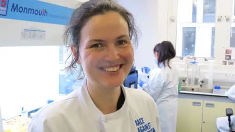 James Gallagher Scientist Dr Claire Durrant smiles. She has tied back hair and looks to the camera in her laboratory. She is wearing a white lab coat with the words "Race Against" clearly written and the word dementia can be inferred. There are people at work behind her.