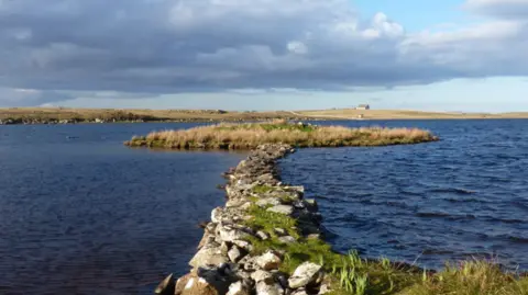 Richard Law The causeway is a narrow, rocky path through the loch to a low, circular grass covered artificial island.