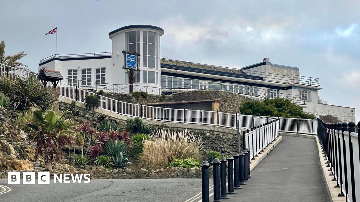 An exterior view of the art deco Winter Gardens building. It is a white building constructed of various shapes, with dirt on some walls. It sits on the cliff edge with a long path leading up to it.