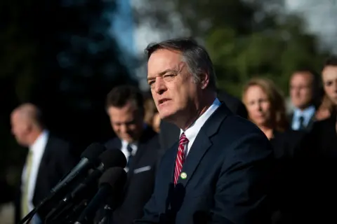 Man with grey hair stands in front of a lecturn, wearing a blue suit, and a red tie. In the background are the blurred 