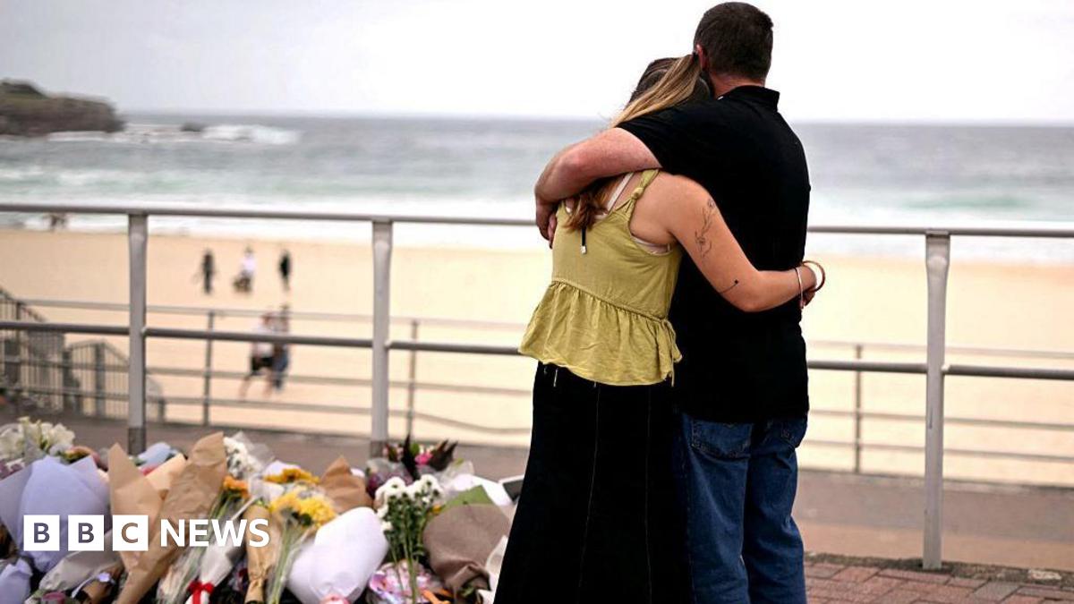 Mourners embrace near tributes piled together in memory of the victims of a shooting at Bondi Beach. A woman wearing a yellow top and black skirt hugs a man wearing a black polo and blue jeans as they look at a pile of flowers. In the background Bondi beach can be seen with people walking on the sand and sea in the distance