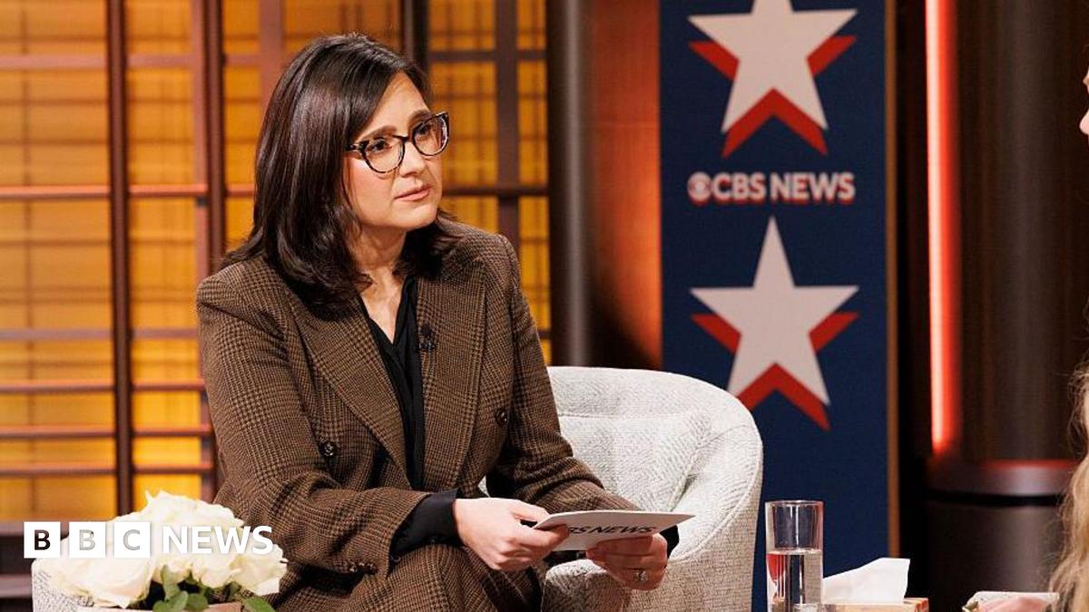 A woman wearing a suit and glasses sits in front of a sign reading CBS News.