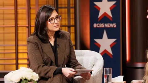 CBS News via Getty Images A woman wearing a suit and glasses sits in front of a sign reading CBS News.