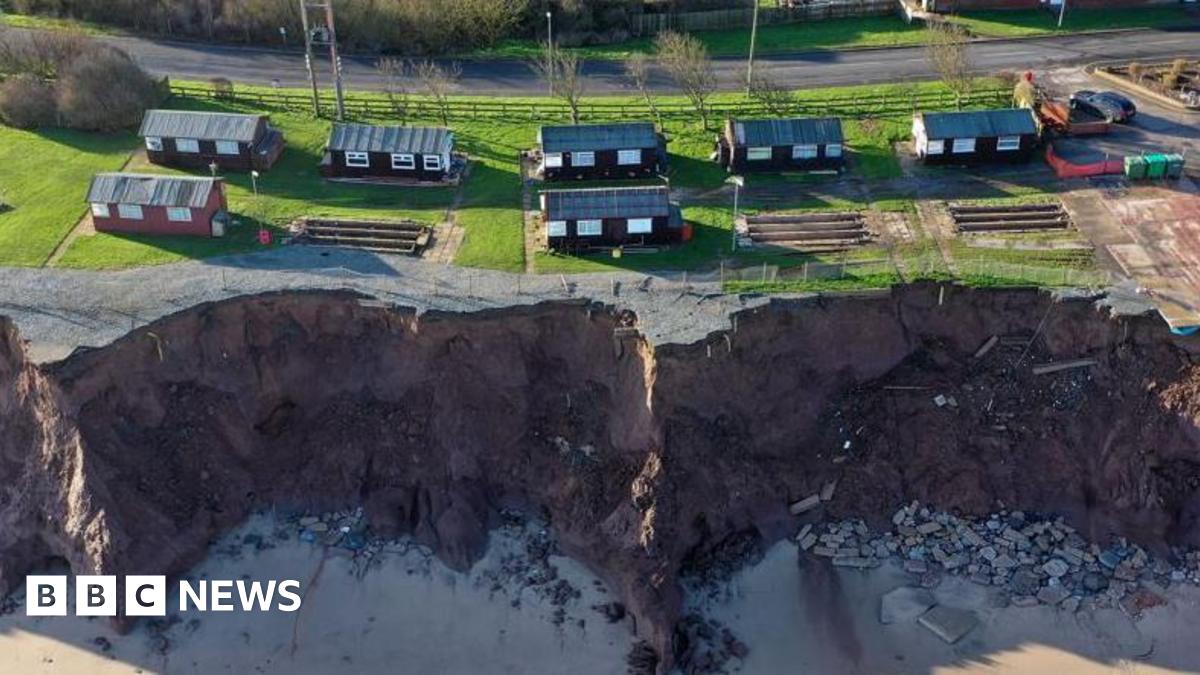 A bird's eye view of dark red holiday chalets dotted along a coastline. The coast is eroding with bits of rock seen to the right of the photo on the sand below, with other items fallen half way down the cliff.