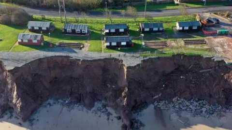 Christopher Furlong/Getty Images A bird's eye view of dark red holiday chalets dotted along a coastline. The coast is eroding with bits of rock seen to the right of the photo on the sand below, with other items fallen half way down the cliff.