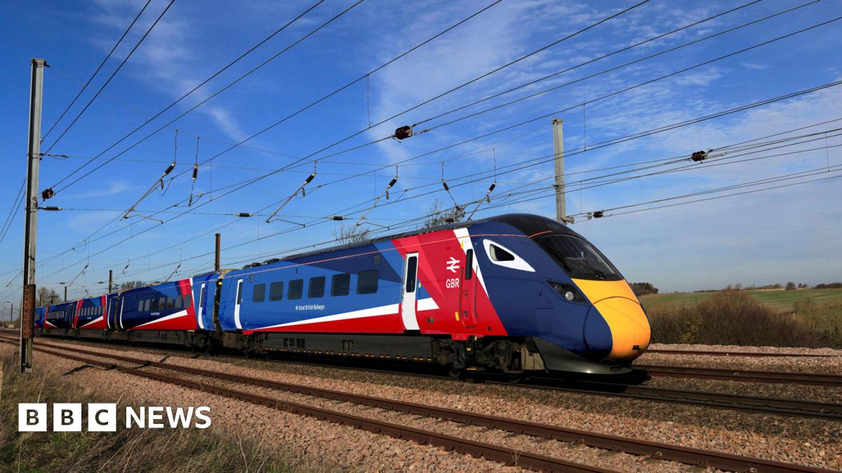 A train with the new red, white and blue branding - it is on a track somewhere in the countryside, with cabling overhead, a blue sky behind it, and fields stretching into the distance.