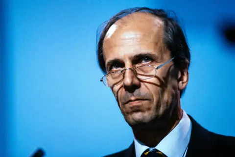 Getty Images Norman Tebbit is wearing a dark pinstripe suit with a white dress shirt and a patterned tie featuring diagonal stripes in yellow and black. The background is a solid blue colour, and part of a microphone is visible on the left side of the image, suggesting a formal speaking or presentation setting.