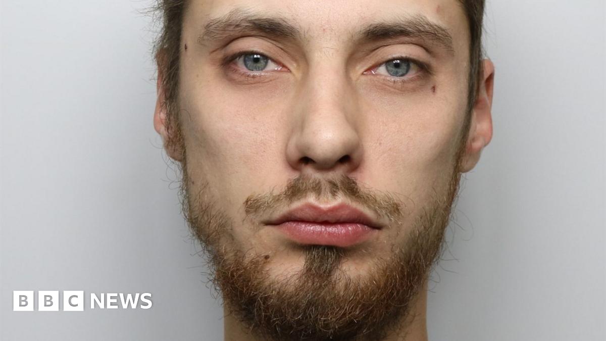 A man with short brown hair and brown or ginger facial hair and blue eyes. It is a custody photo and the background is grey.