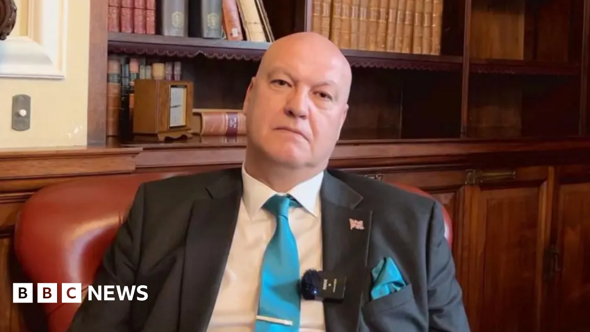 Ian Cooper sits in a maroon leather chair, with wooden panelling and a recessed bookcase behind him. He is wearing a white shirt, dark grey suit jacket, with a union flag pin badge and a clip microphone on the lapel, and a teal blue tie. He is looking into the camera with a serious expression.