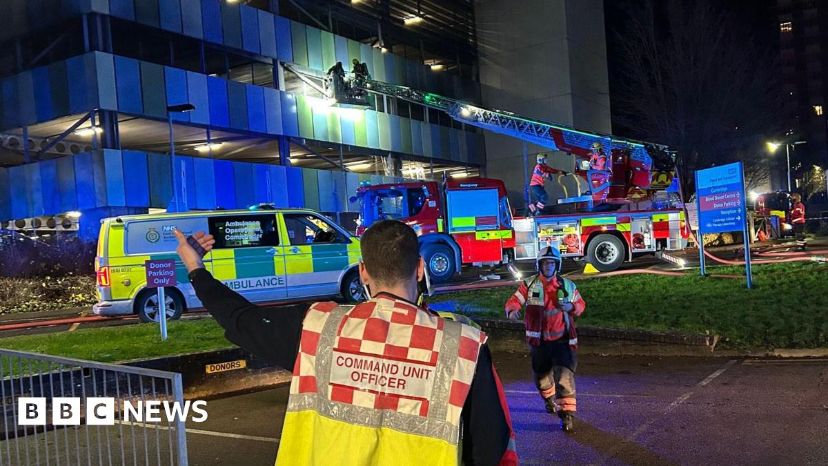 In the foreground, a person has their back turned to the camera, and their vest reads COMMAND UNIT OFFICER. In the background is a fire engine with a cherry picker extending to a modern multi-storey car park building. It is dark. There is an ambulance vehicle as well.