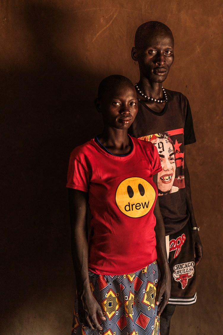 A woman and a man stand together looking at the camera in a dark room, casting a large shadow on the wall behind them.