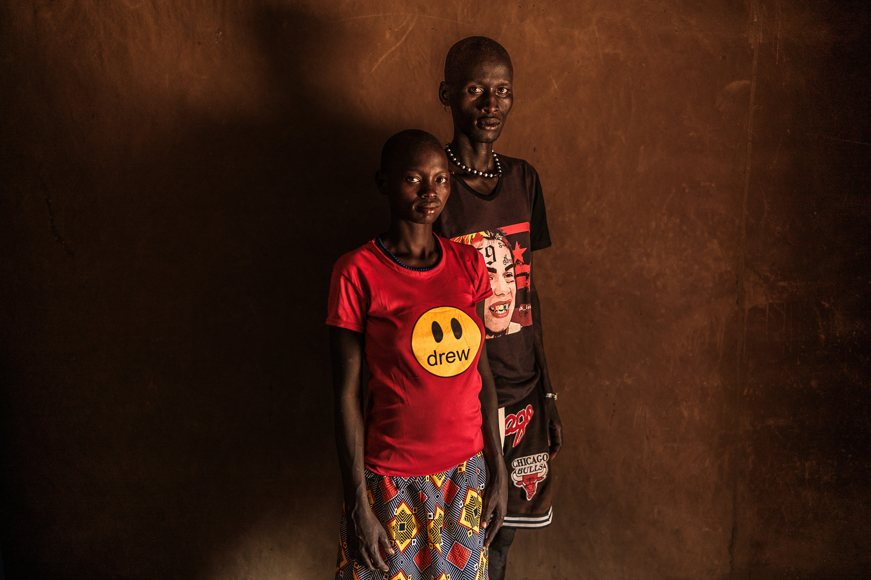 A woman and a man stand together looking at the camera in a dark room, casting a large shadow on the wall behind them.