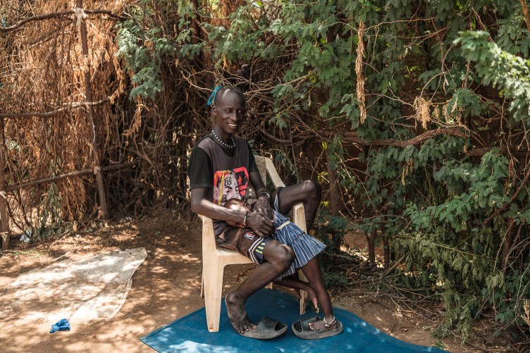 A man sits on a plastic chair. A small girl stands next to him, resting her head in his lap as he holds her. They are outdoors, with a dirt floor, surrounded by scrub brush and simple fencing.