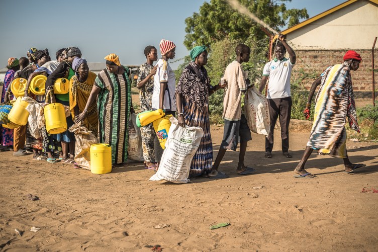 A man lifts a rope for a line of people to pass underneath. People in the crowd hold jerricans or sacks.