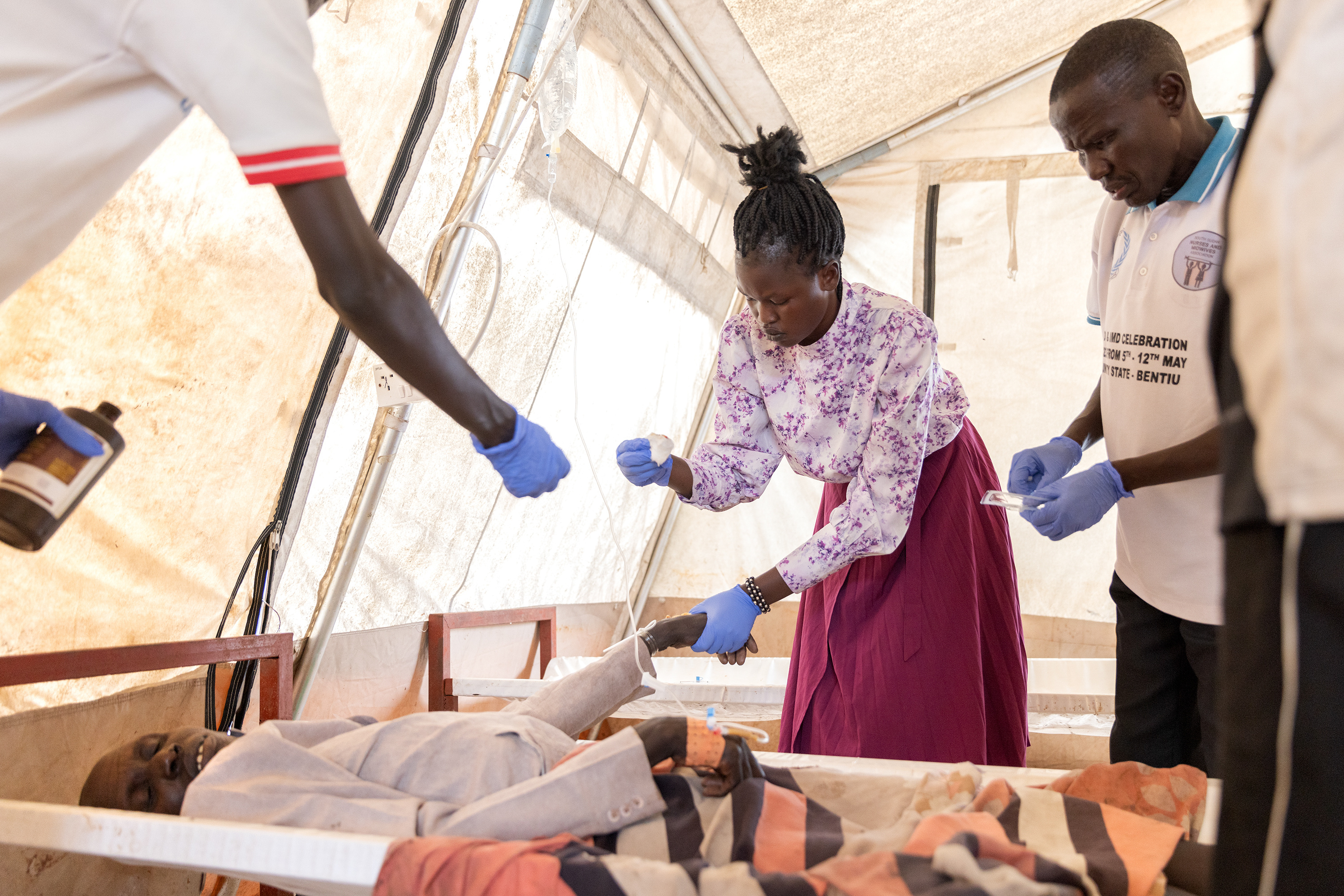 A chaotic scene in which a patient is lying on a hospital bed in a tent, apparently unconscious, a woman is holding his hand and cleaning an injection site for an IV, and two other people are leaning in to offer her supplies.