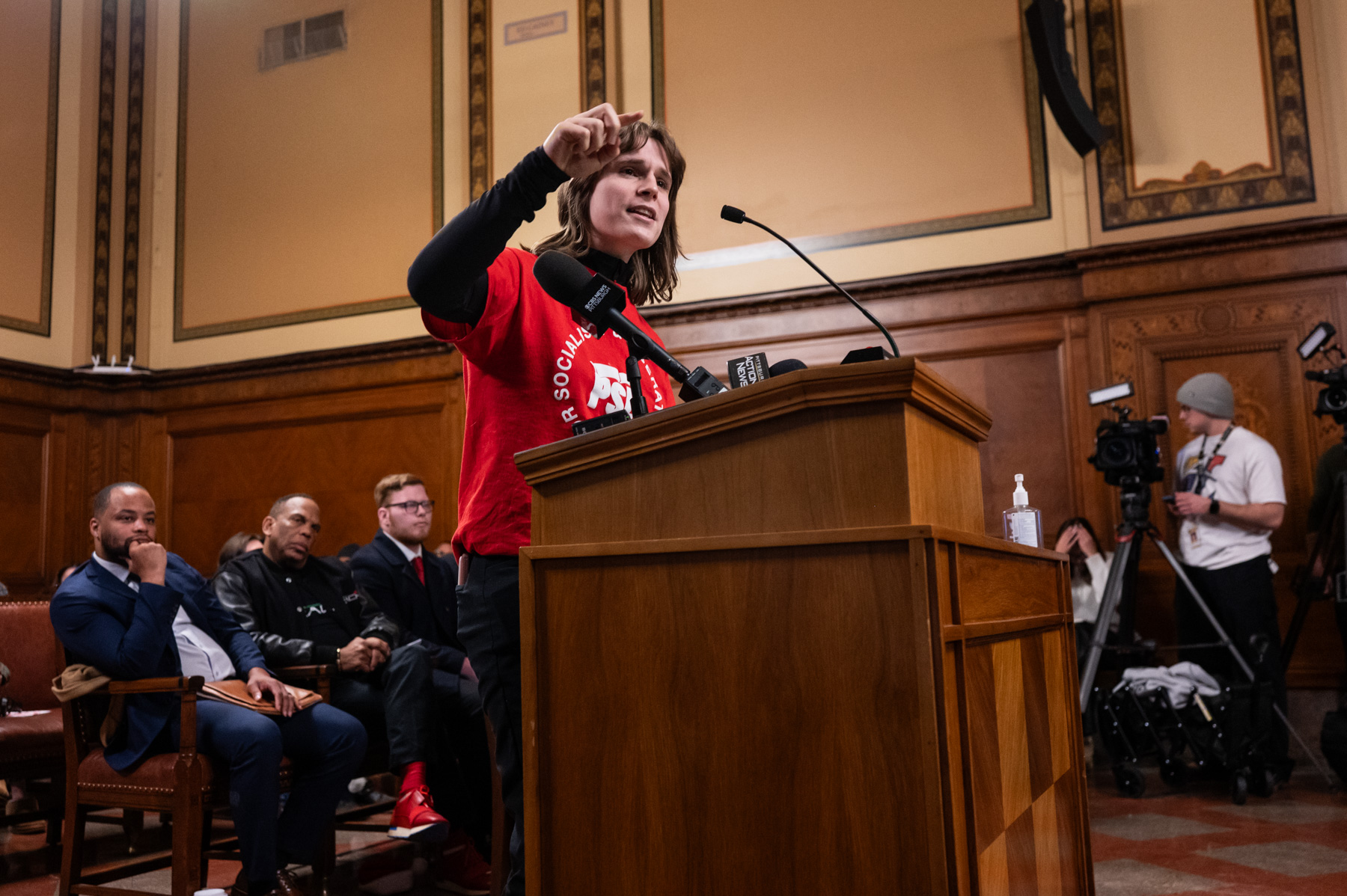 A person in a red shirt speaks at a podium in a formal room, with seated attendees and photographers visible in the background.