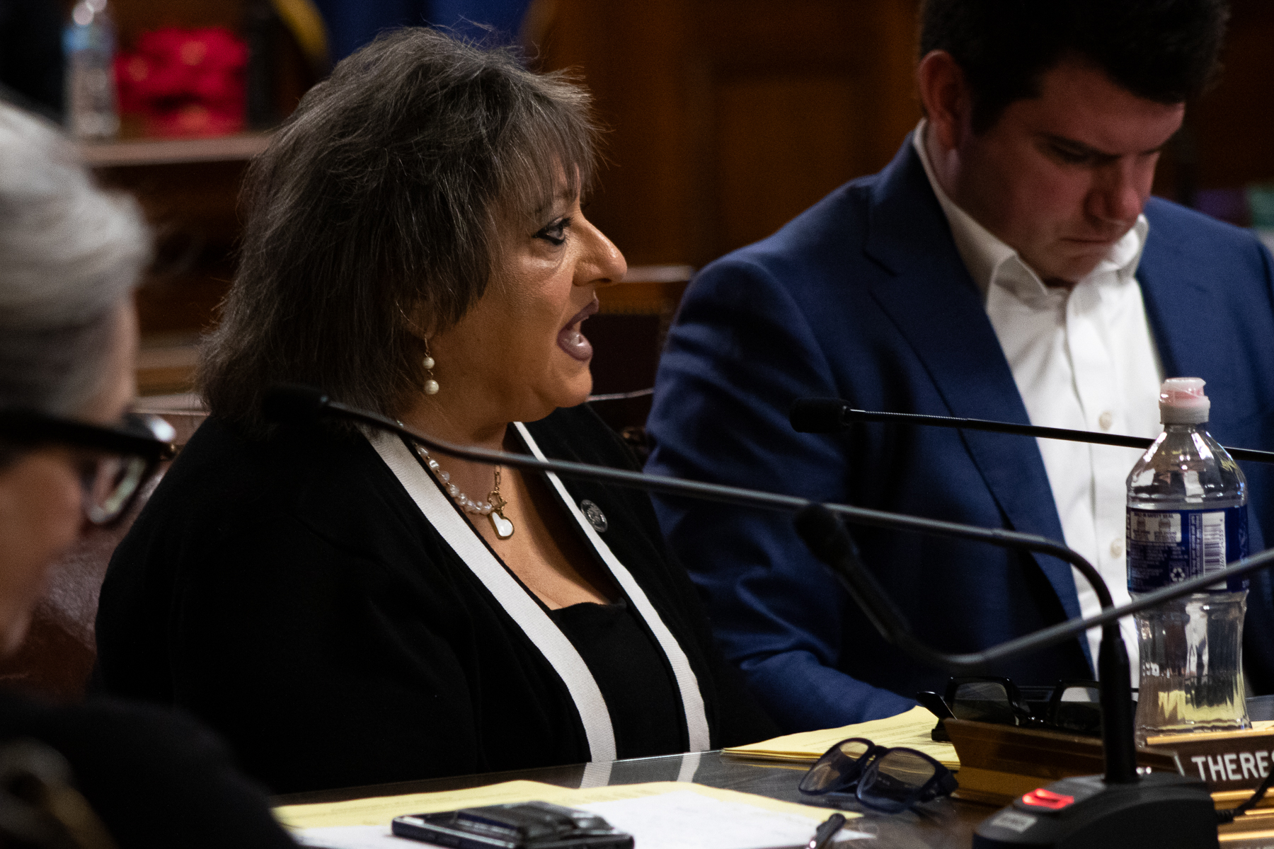 A woman speaks into a microphone at a meeting while a man beside her reads a document. Both are seated at a table with nameplates, papers, and a water bottle.