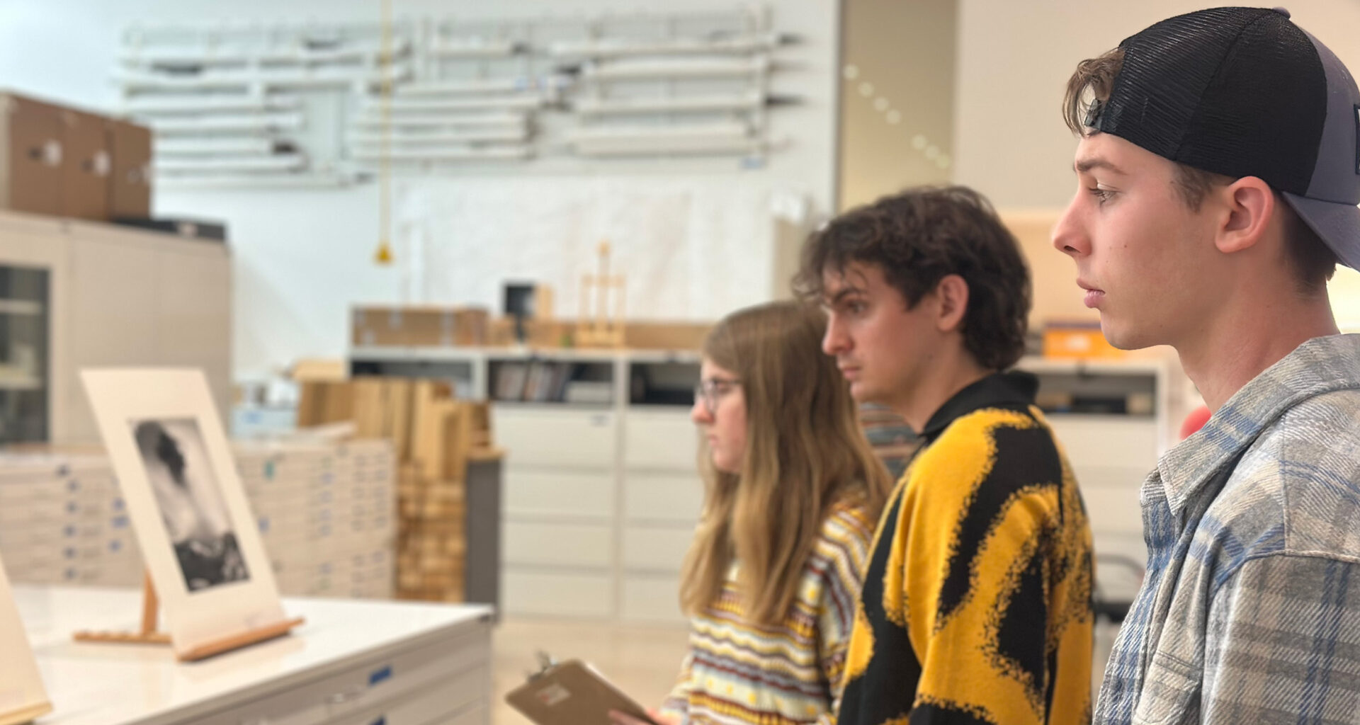 Three people standing indoors in front of a counter, observing framed artwork on display; shelves and boxes are visible in the background.