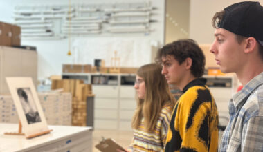 Three people standing indoors in front of a counter, observing framed artwork on display; shelves and boxes are visible in the background.