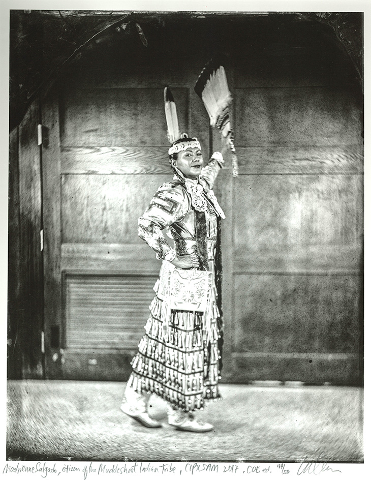 Person wearing a traditional jingle dress with metal cones, standing indoors in front of wooden double doors, holding two feather fans raised in one hand.