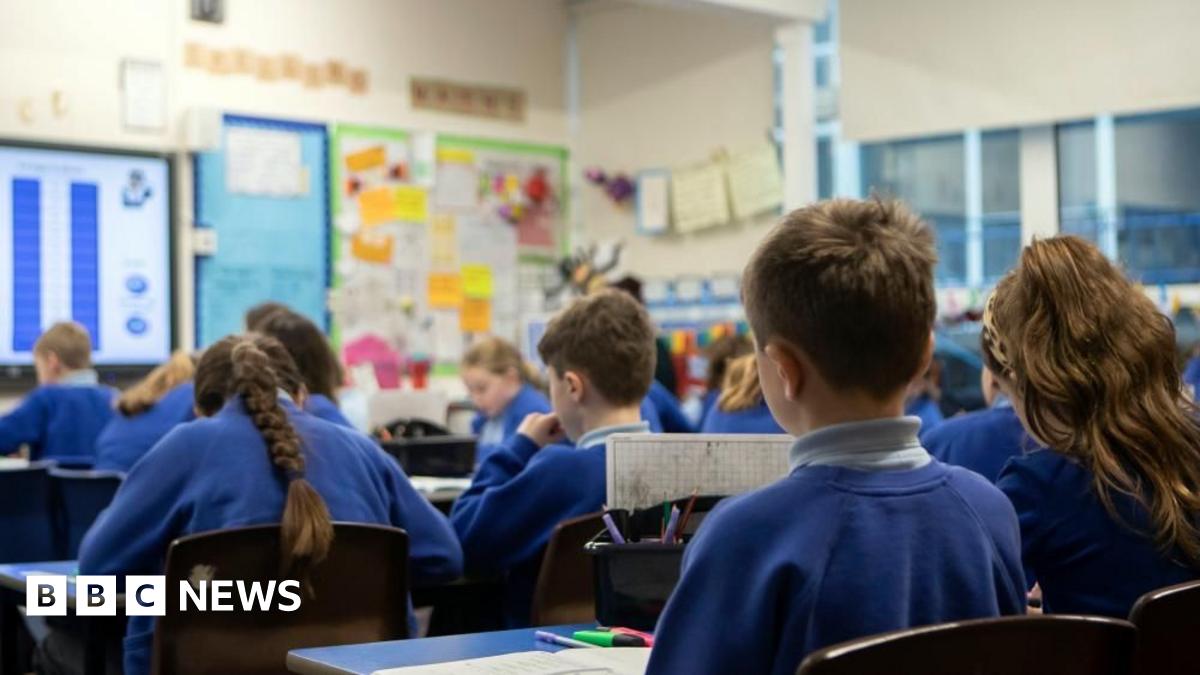 A group of year five pupils sat down facing the front of a classroom. The students are wearing blue jumpers and blue polo shirts and none of their faces are visible.