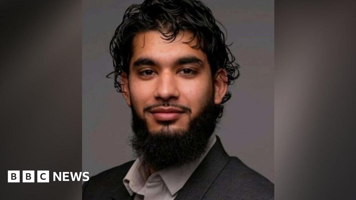 Adam Khan: a man who is sitting looking at the camera and smiling. He has black hair and black facial hair. He is wearing a grey suit jacket and light grey shirt.