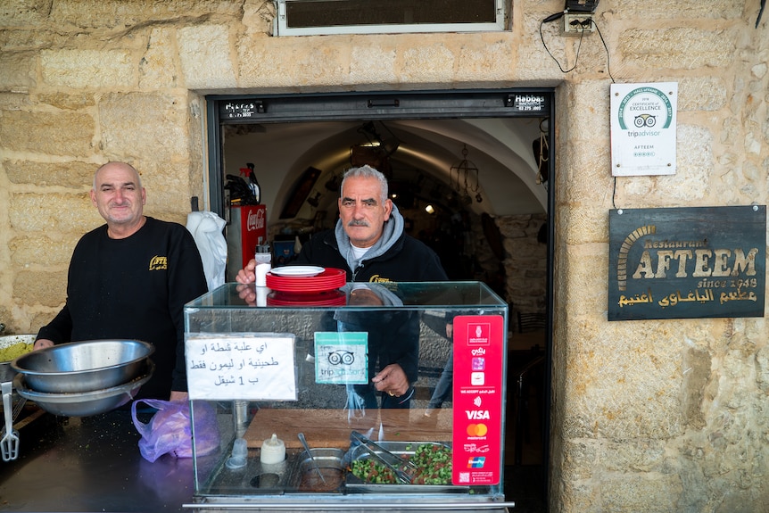 A man stands a small a glass box serving food, in front of a door way with a sign on the wall reading Afteem
