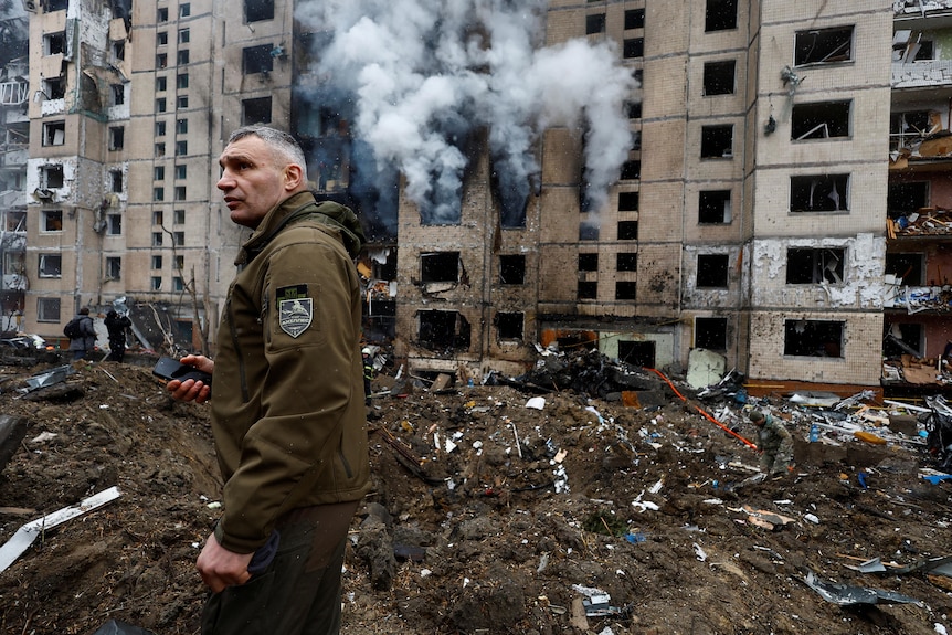 Man with army green jacket walks amid building smoke and debris 