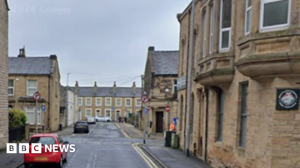 A red car is parked on Nelson Street with stone terraced houses and businesses on both sides of the road