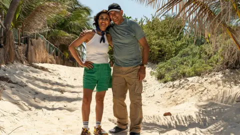 StudioLambert/BBC Anita Rani and dad Bal stand on a sandy beach smiling at the beginning of their journey.