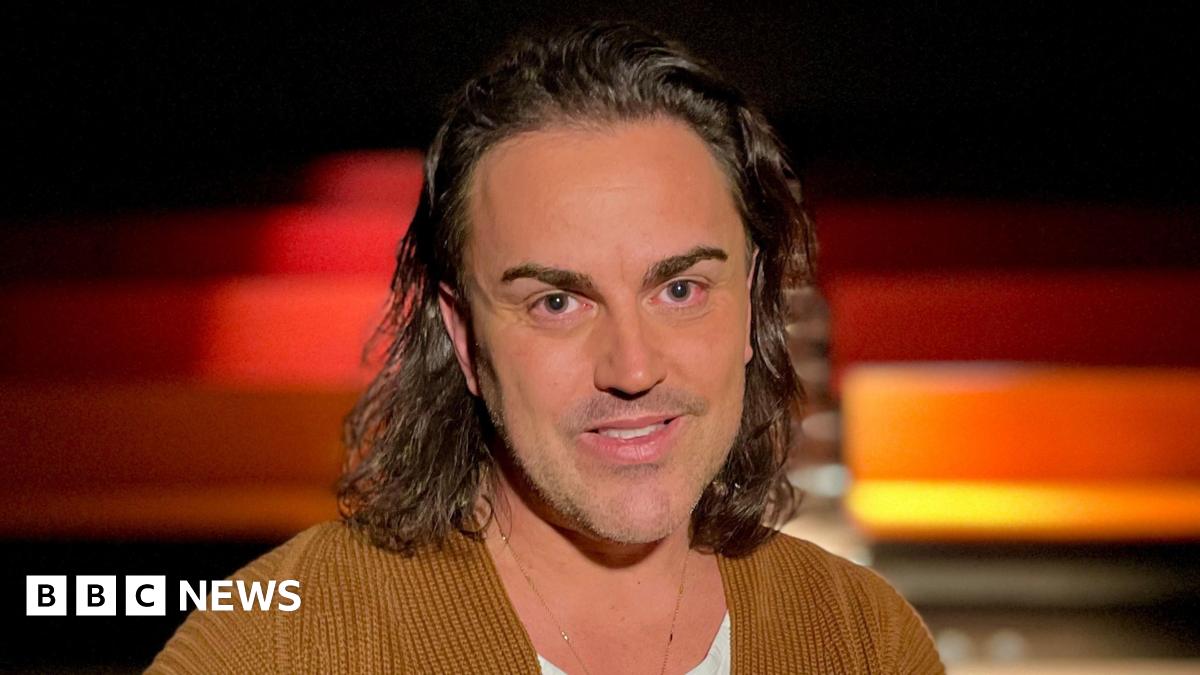 Stephen Crocker is in the middle of the frame. He has shoulder-length brown curly hair and is wearing a white T-shirt and a brown jacket. Behind him are rows of seats.