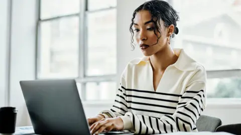 Getty Images Young black woman with black curly hair tied up sitting in an office wearing a cream and black striped jumper typing on a laptop