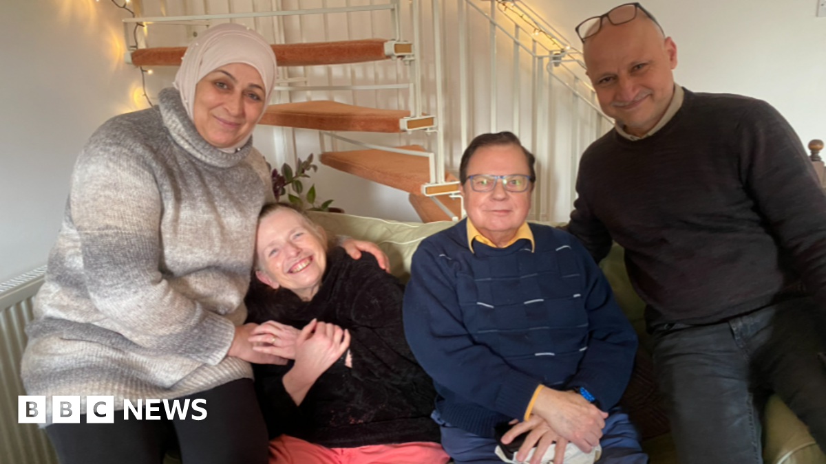 Pictured is Bushra, Margaret, George and Khaled all sat on a white leather sofa in the living room. Bushra is wearing a nude headscarf with pearl embellishments attached on it, a high neck wool-like jumper that is grey, brown and beige. Margaret is wearing a black jumper and pink trousers. She has her head laid on Bushra and is smiling. George wears a yellow collar shirt underneath a blue jumper and has slicked-back brown hair. Khaled has his arm wrapped behind George's shoulder and is smiling. He wears a dark brown jumper with a beige collar shirt underneath and dark blue jeans with his black framed glasses sitting on the top of his head.
