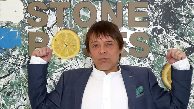 Mani in a suit standing in front of an artwork of the Stone Roses first album