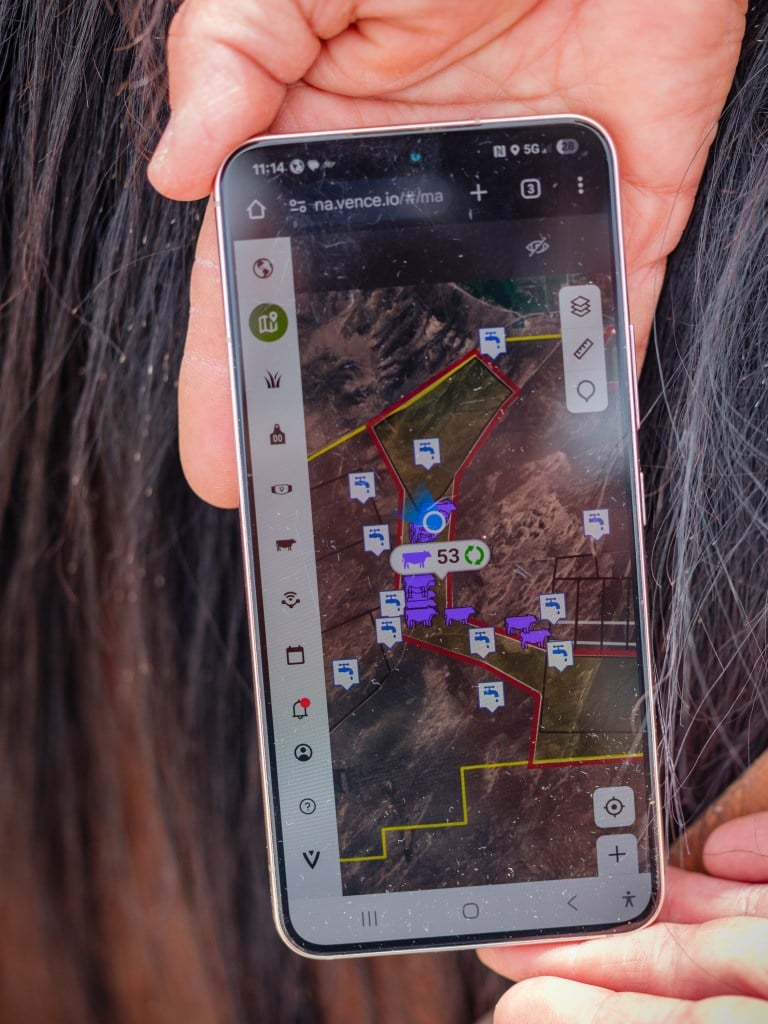 Hands hold out a mobile phone, displaying virtual fencing and water sources for cattle grazing over a satellite view of the area. The screen shows purple cow icons confined within red and yellow lines, as well as water faucet icons.
