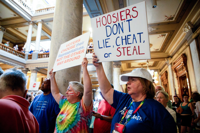 People rally inside of a state house, a person holds a sign that says "Hoosiers don't lie, cheat or steal" while another holds a sign that reads "Voters choose their representatives, not the other way around"