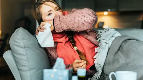 Getty Images A woman with flushed cheeks lies on a couch covered in blankets. She covers her mouth, apparently coughing, and has a tissue in her hand. She has long blonde hair in a plait and is wearing a pink jumper and a coral scarf. In the foreground of the shot is a box of tissues, a mug and two medicine bottles.