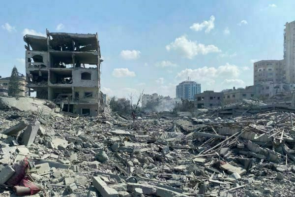 A multi-story building with its upper floors destroyed stands amidst vast piles of grey concrete rubble. In the midground, smoke rises from more debris, with damaged structures and a modern blue skyscraper visible in the distance under a cloudy sky.
