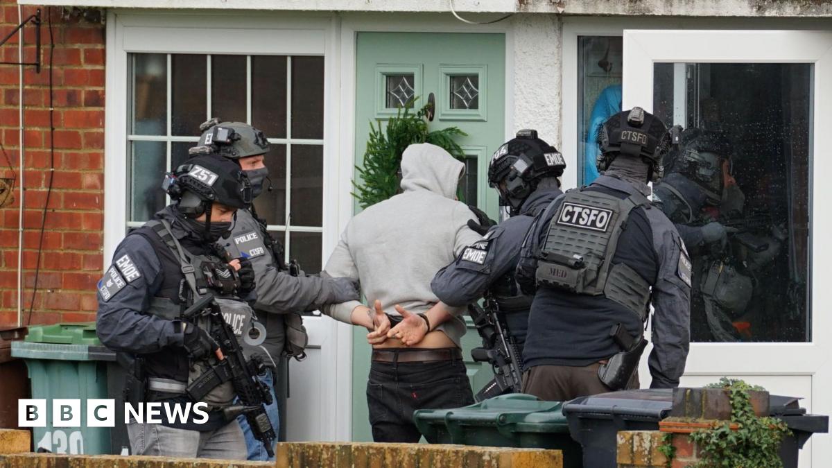 A man in a grey hoodie facing away from the camera, with handcuffs on behind his back. He has armed police officers either side of him, with the letters ‘CTSFO', 'EMOE' and 'POLICE' on their uniforms.